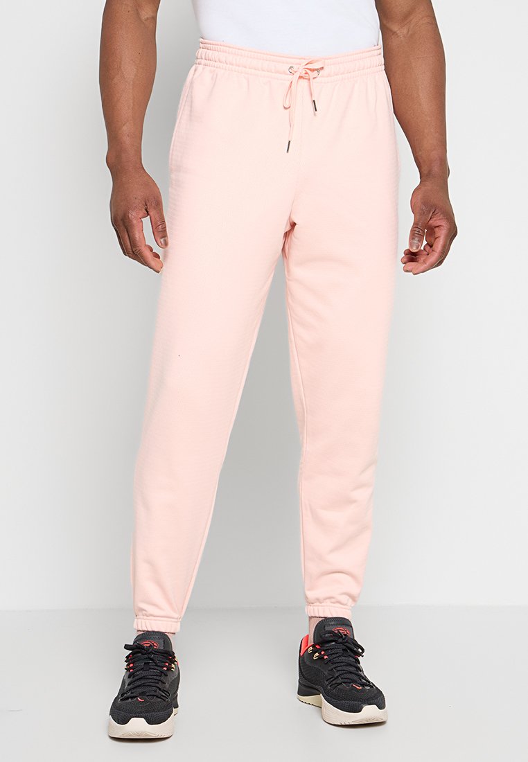 Mann trägt hellrosa Sweatpants mit Kordelzug und schwarze Sportschuhe, steht vor einem schlichten Hintergrund.