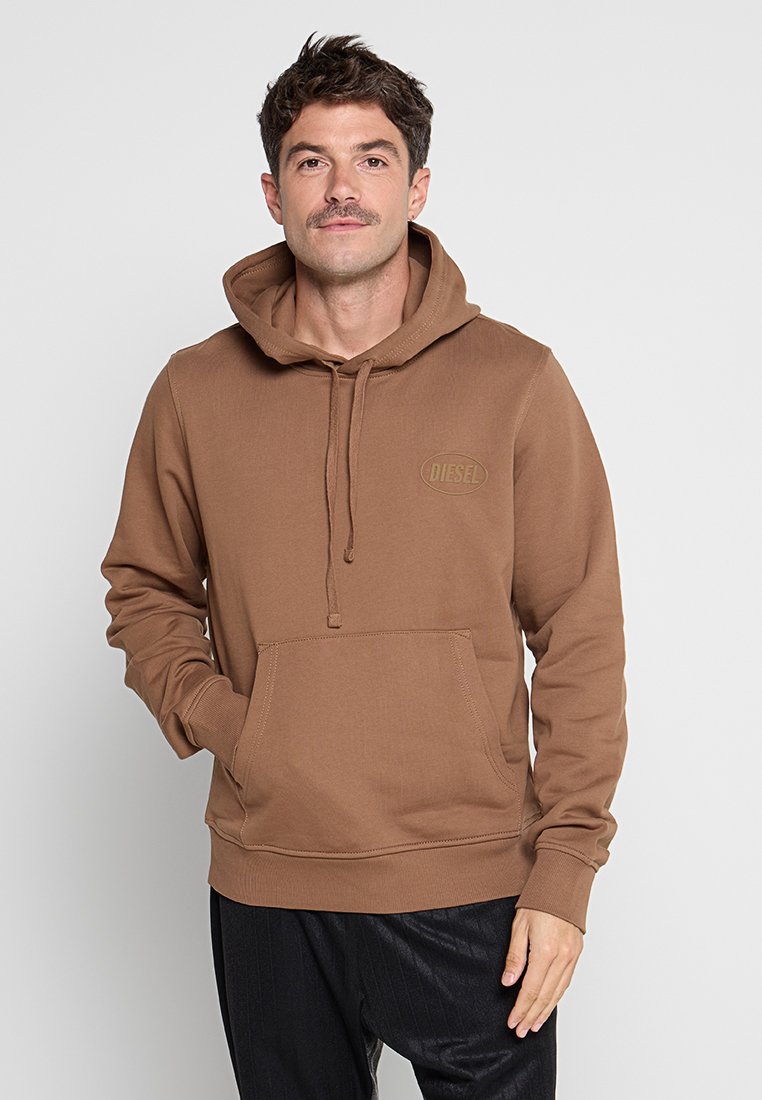 Mann mit Schnurrbart, der einen braunen Diesel-Hoodie mit Fronttasche und schwarze Hosen trägt, steht vor einem schlichten, hellen Hintergrund.
