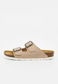 Beige Wildleder-Pantoletten mit zwei verstellbaren Metallschnallenriemen und einem Korkfußbett auf einer weißen strukturierten Sohle.