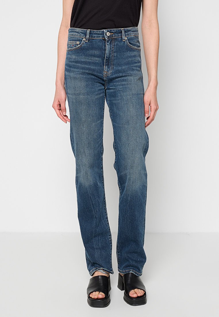 Person trägt blaue Jeans mit mittlerem Bund und geradem Bein, schwarzes ärmelloses Top sowie schwarze offene Sandalen mit Absatz und steht vor einem schlichten Hintergrund.