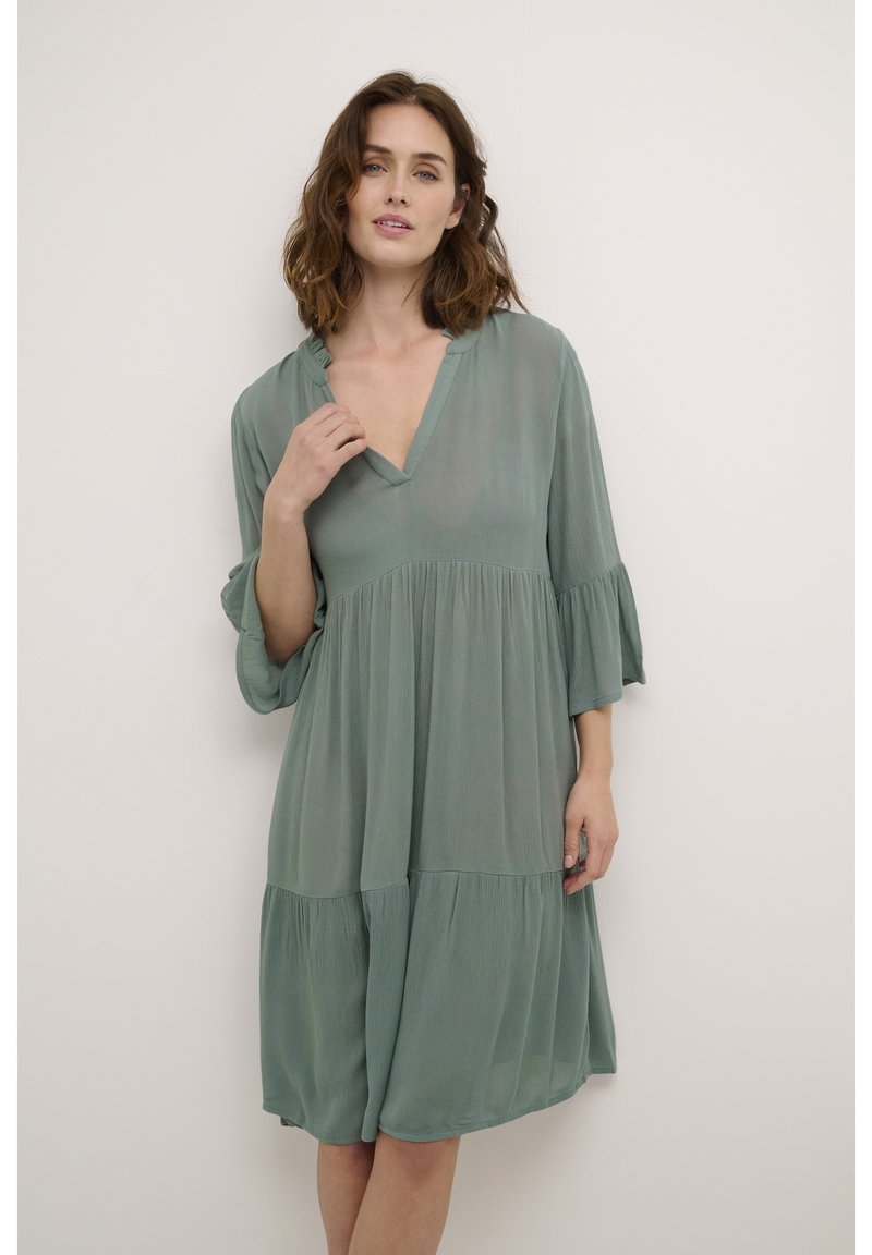 Helles grünes, lässig geschnittenes Kleid mit V-Ausschnitt und 3/4-Bellärmeln. Hat ein gestuftes Design und eine weiche, fließende Textur.