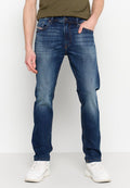 Mann in blauen, ausgewaschenen Slim-Fit-Jeans, weißen Sneakers und einem olivgrünen T-Shirt, der mit einer Hand in der Tasche steht.