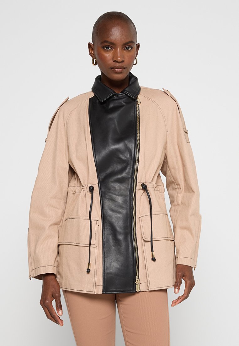 Beige Stoffjacke mit einem schwarzen Leder-Vorderteil, ausgestattet mit zwei Fronttaschen, einem verstellbaren Bund und Schulterklappen. Mit Reißverschluss.