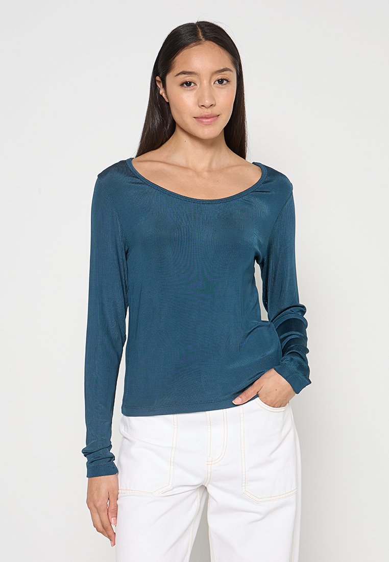 Türkises Langarmshirt mit rundem Ausschnitt und geripptem Muster, kombiniert mit weißen Hosen, die sichtbare Nähte aufweisen.