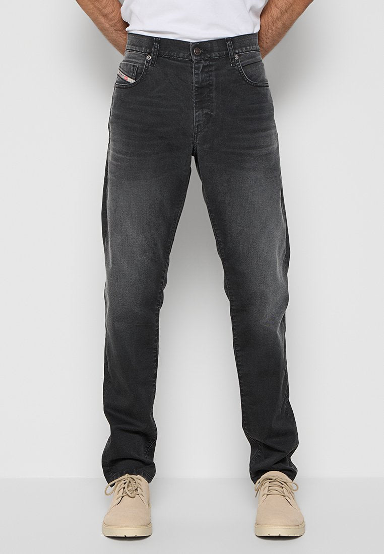 Mann in verblichenen schwarzen gerade geschnittenen Jeans und beigen Schnürschuhen, stehend vor einem schlichten hellen Hintergrund.
