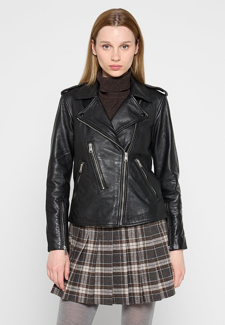 Schwarze Lederbikerjacke mit Reißverschlusstaschen und Schulterklappen, mit tailliertem Schnitt. Tragen über einem braunen Rollkragenpulli und einem karierten Rock.