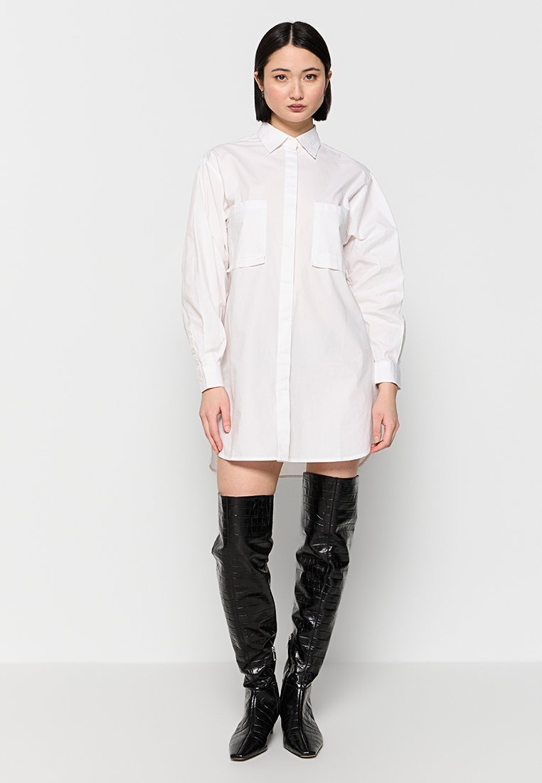 Weißes Hemdkleid mit zwei Brusttaschen, langen Ärmeln und geradem Schnitt, kombiniert mit schwarzen, glänzenden Overkneestiefeln.