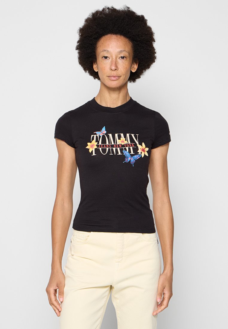 Schwarzes, tailliertes T-Shirt mit einem bunten "TOMMY JEANS NEW YORK"-Logo, versehen mit floralen und Schmetterlingsgrafiken. Kurze Ärmel und gerippte Textur.