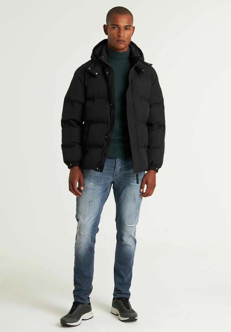 Schwarze Pufferjacke mit Kapuze, ausgestattet mit einem Reißverschluss und elastischen Bündchen, kombiniert mit hellblauen, ausgewaschenen Jeans und dunklen Sneakers.
