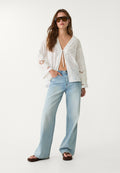 Weiße Spitzenbluse mit Glockenärmeln, vorne zum Binden und Ausschnitten, kombiniert mit hellblauen weit geschnittenen Jeans und braunen Flip-Flops.