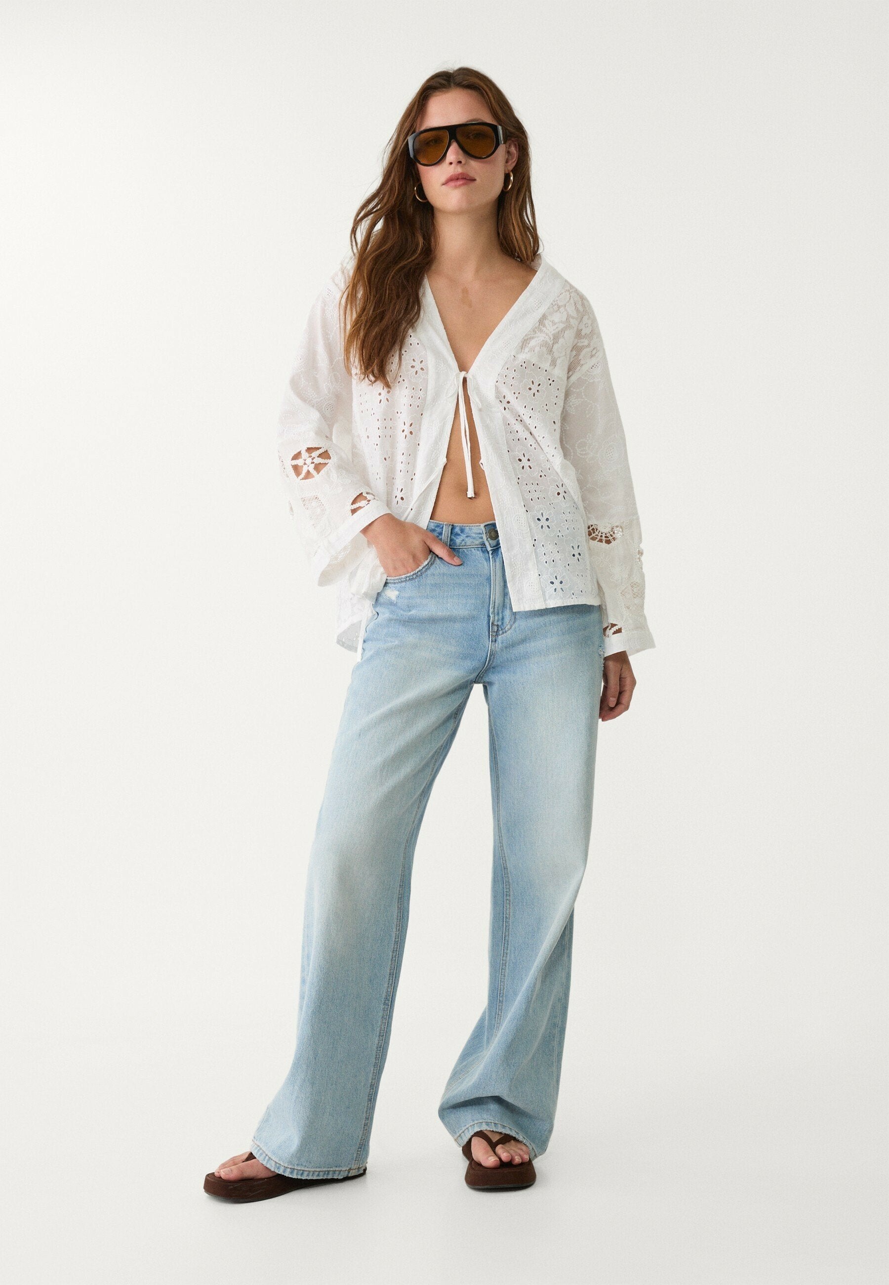 Weiße Spitzenbluse mit Glockenärmeln, vorne zum Binden und Ausschnitten, kombiniert mit hellblauen weit geschnittenen Jeans und braunen Flip-Flops.