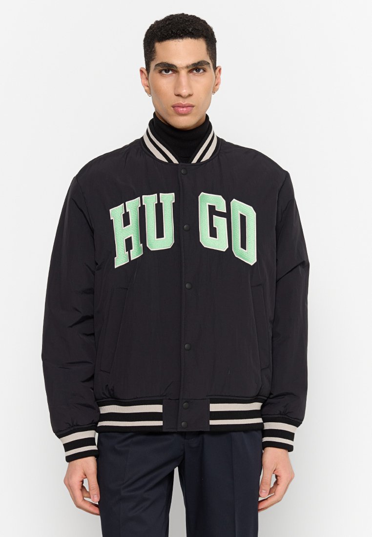 Schwarze Bomberjacke mit weißen und hellgrünen „HUGO“-Buchstaben, gerippten gestreiften Bündchen und Saum sowie Druckknopfverschluss. Glatte Stofftextur.