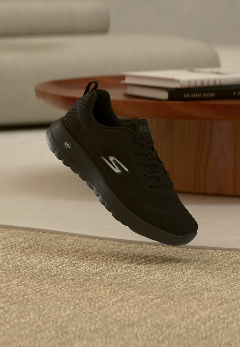 Schwarzer Skechers-Sneaker mit weißem Logo schwebt über beigem Teppich neben einem hölzernen Couchtisch mit gestapelten Büchern in einem Wohnzimmer.