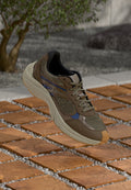 Brauner Sportschuh aus Mesh mit grauen Akzenten, einer strukturierten Oberseite, dicker Sohle und einem blauen Streifen. Präsentiert auf hölzernen Fliesen mit Kies.