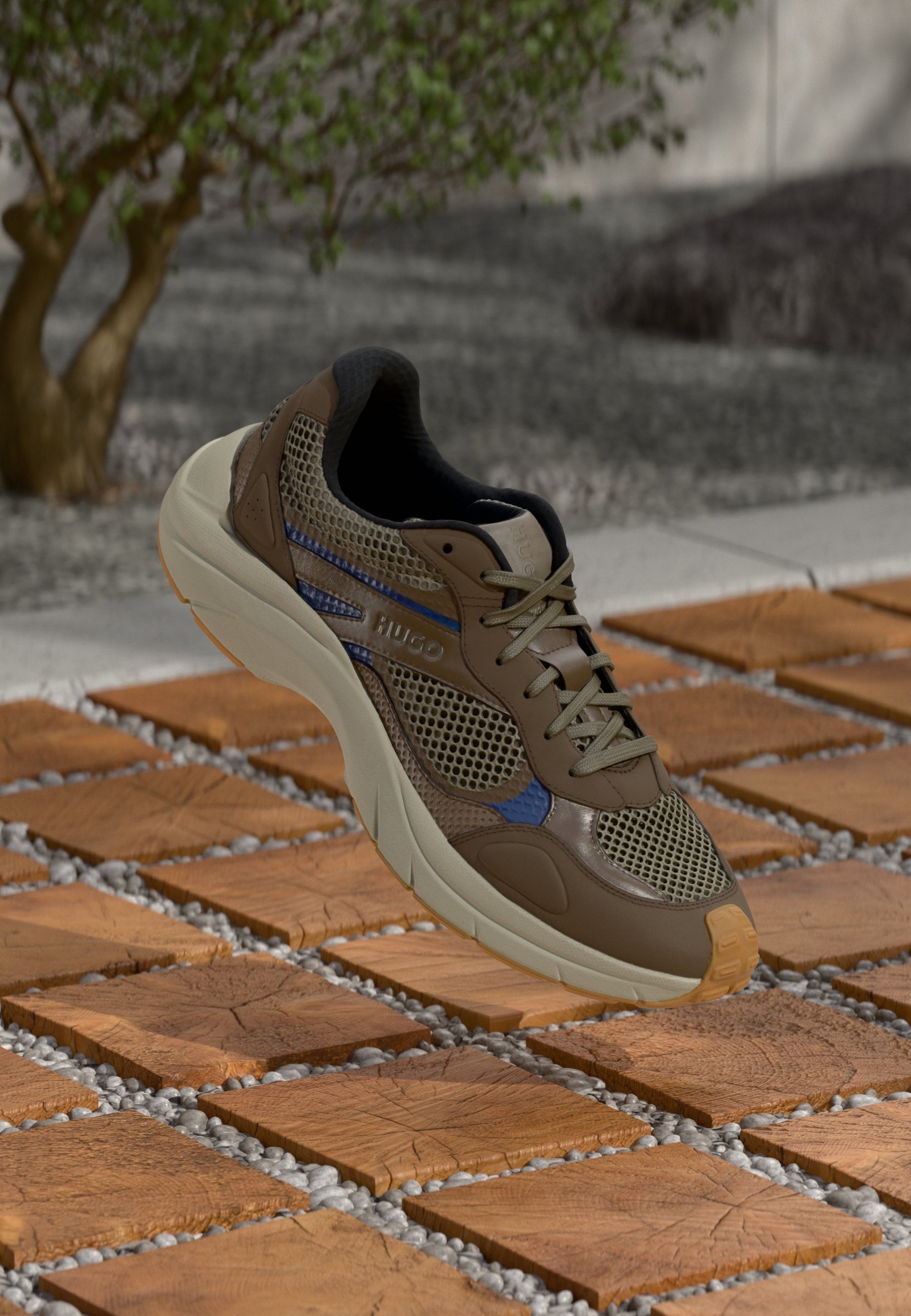 Brauner Sportschuh aus Mesh mit grauen Akzenten, einer strukturierten Oberseite, dicker Sohle und einem blauen Streifen. Präsentiert auf hölzernen Fliesen mit Kies.