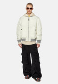 Weiße gepolsterte Jacke mit grauen Akzenten, über einem beigen Hoodie getragen. Schwarze Cargo-Hosen mit mehreren Taschen. Kombiniert mit dunklen Sonnenbrillen und gemusterten Schuhen.