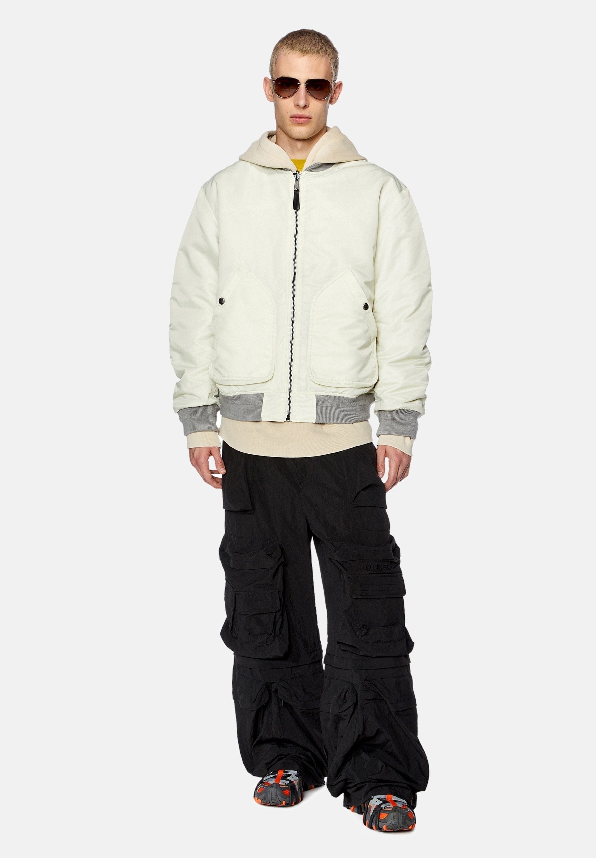 Weiße gepolsterte Jacke mit grauen Akzenten, über einem beigen Hoodie getragen. Schwarze Cargo-Hosen mit mehreren Taschen. Kombiniert mit dunklen Sonnenbrillen und gemusterten Schuhen.