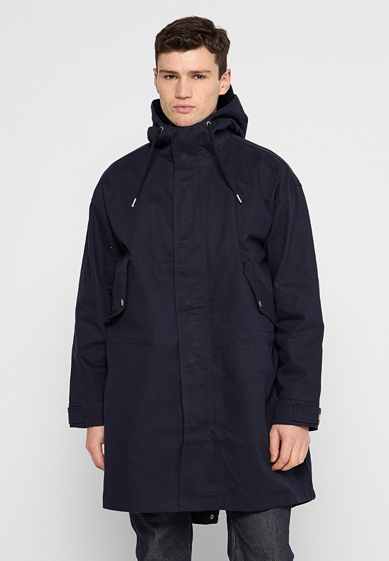 Ein marineblauer langer Mantel aus Stoff, mit hohem Kragen, Kordelzugkapuze, zwei vorderen Taschen und Knopfverschlüssen.