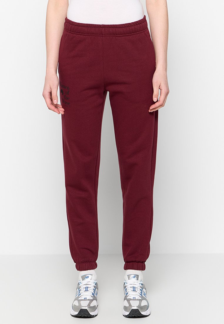 Burgunderfarbene Sweatpants aus Baumwollmix mit elastischem Bund, elastischen Bündchen und einem bedruckten Grafikdesign auf der Vorderseite. Kombiniert mit grauen Sneakers.