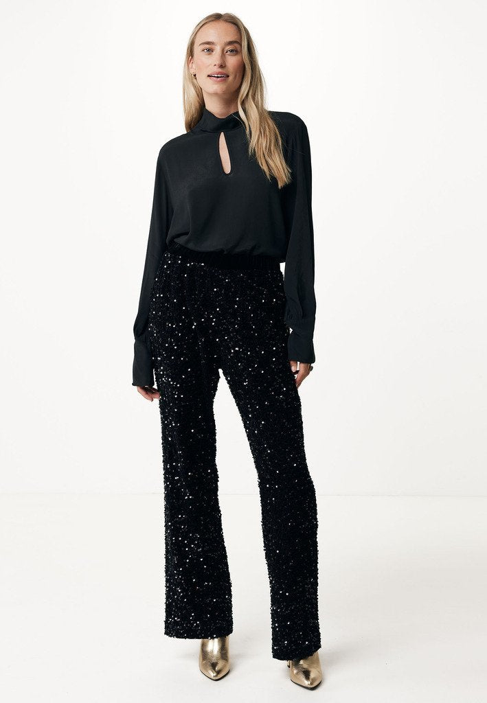 Schwarze satinierte Bluse mit Schlüsselloch-Ausschnitt, kombiniert mit schwarzen Pailletten-Hosen mit weiter Beinsilhouette und metallic-goldenen High Heels. Glatte Textur, glänzende Akzente.