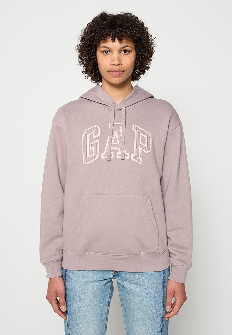 Junge Person mit lockigem Haar, die einen mauvefarbenen GAP-Kapuzenpullover und hellblaue Jeans trägt, vor einem schlichten weißen Hintergrund stehend.
