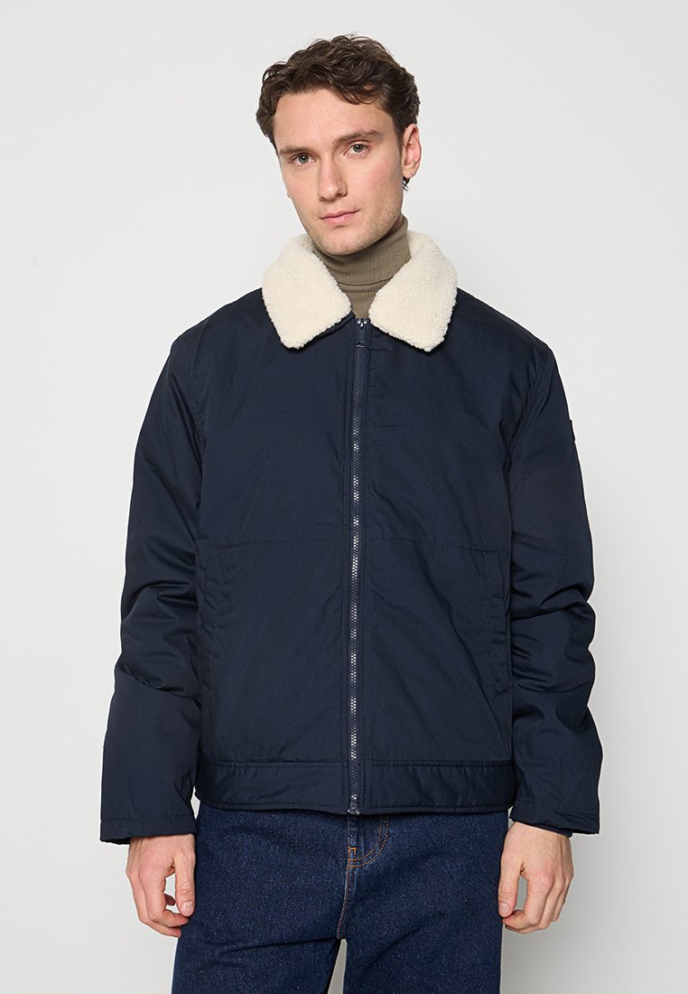Marineblaue isolierte Jacke mit einem flauschigen cremefarbenen Kragen, Reißverschluss und Seitentaschen; zeichnet sich durch eine glatte, weiche Textur aus.