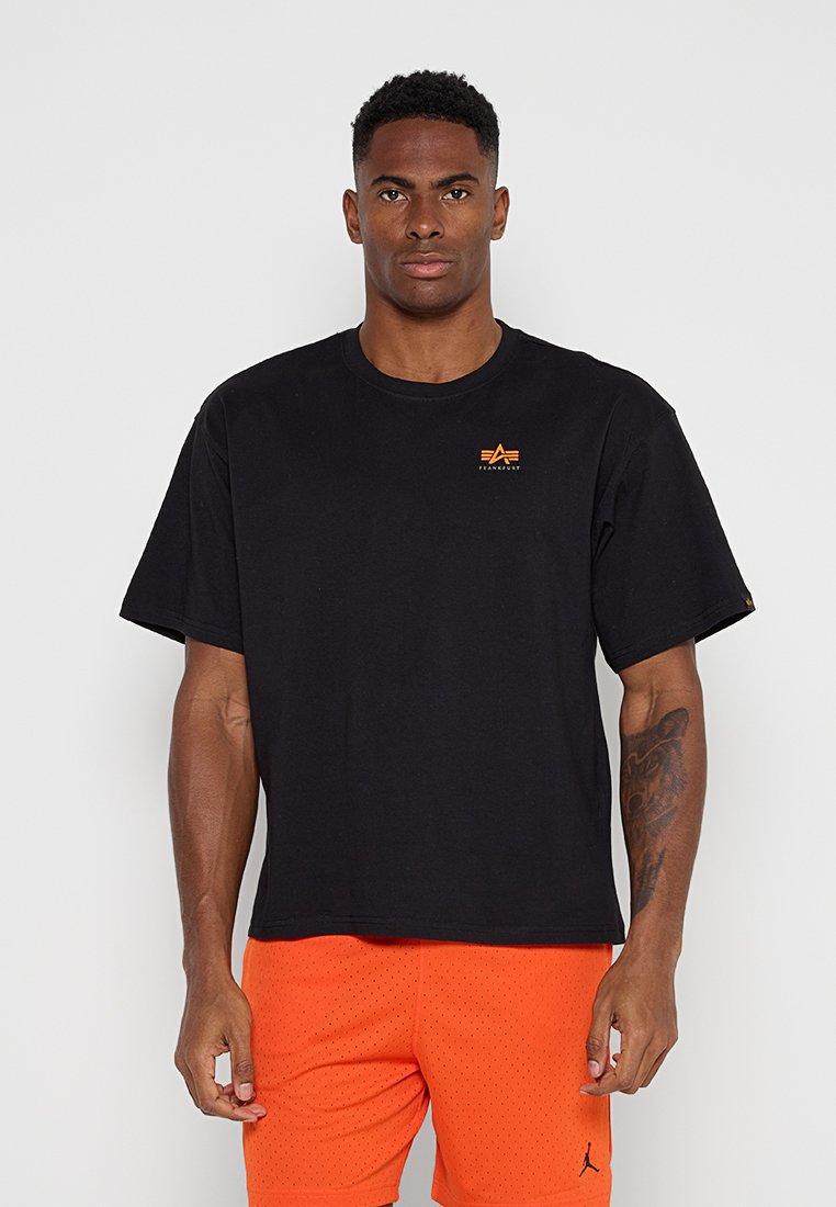 Mann trägt schwarzes kurzärmeliges Hemd mit kleinem orangefarbenem Logo und hellorangene, perforierte Shorts, steht vor einem einfarbigen grauen Hintergrund.