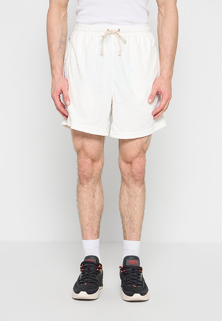 Mann trägt weiße Shorts mit Kordelzug, weiße Socken und schwarze Sportschuhe, steht vor einem einfarbigen weißen Hintergrund.