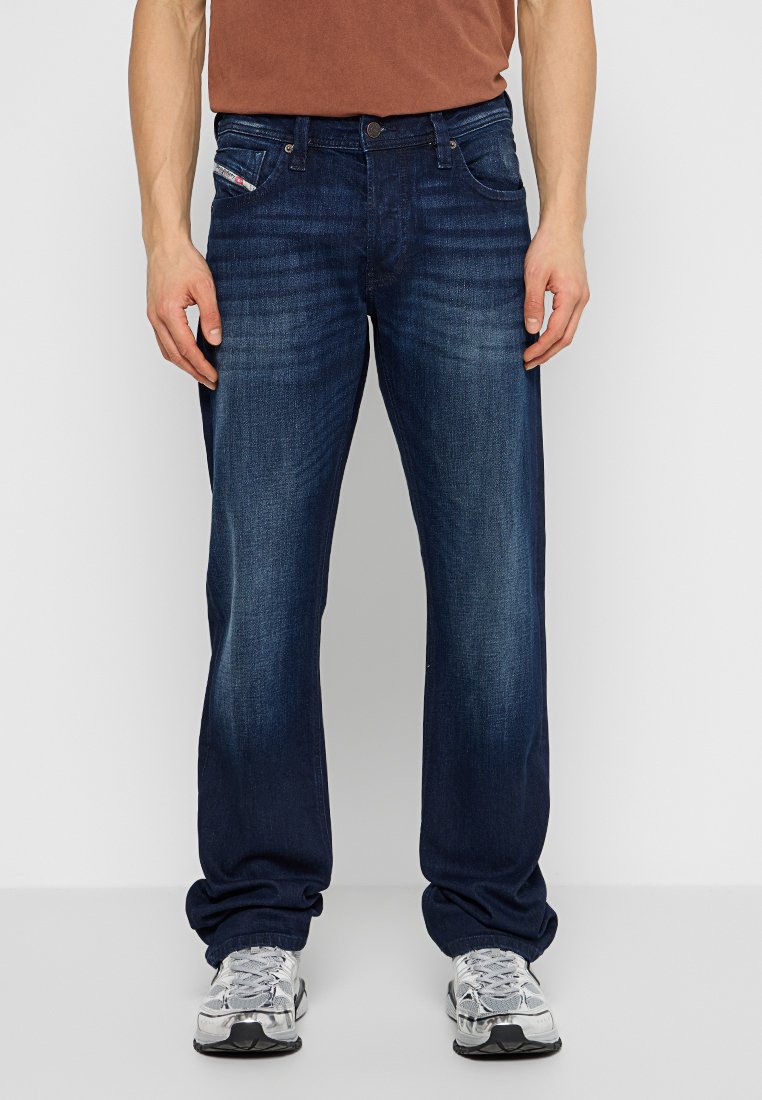 Blaue Jeans aus Denim mit geradem Bein, subtiler Ausbleichung, Fünf-Taschen-Design und silberner Knopfleiste, kombiniert mit einem braunen Hemd.
