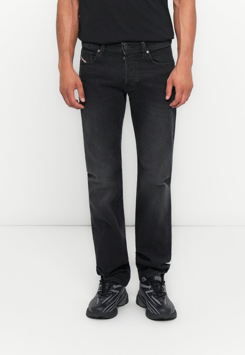 Mann in schwarzen, ausgewaschenen Jeans, schwarzen Sneakers mit grauen Akzenten und einem schwarzen T-Shirt, der vor einem weißen Hintergrund steht.