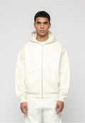 Junger Mann trägt einen elfenbeinfarbenen Hoodie mit Reißverschluss und passende Hose, steht vor einem schlichten hellgrauen Hintergrund.