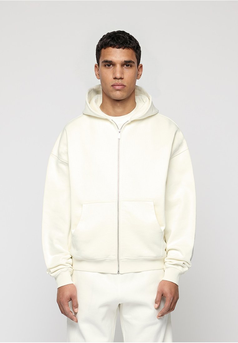Junger Mann trägt einen elfenbeinfarbenen Hoodie mit Reißverschluss und passende Hose, steht vor einem schlichten hellgrauen Hintergrund.