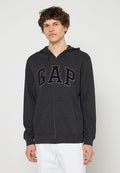 Junger Mann trägt einen dunkelgrauen Kapuzenpullover mit Reißverschluss und den Buchstaben "GAP" sowie weiße Hose vor einem schlichten hellen Hintergrund.