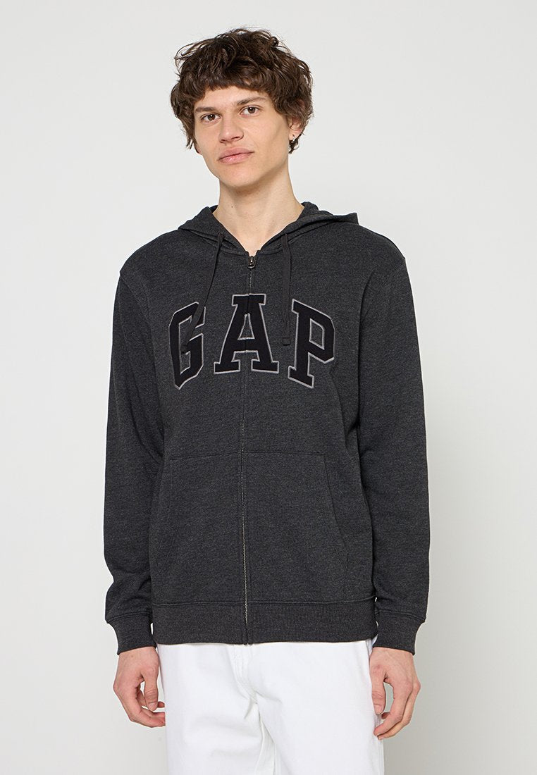 Junger Mann trägt einen dunkelgrauen Kapuzenpullover mit Reißverschluss und den Buchstaben "GAP" sowie weiße Hose vor einem schlichten hellen Hintergrund.