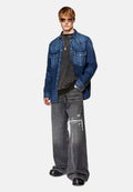 Jeansjacke mit zwei Brusttaschen, kombiniert mit einem dunkelgrauen T-Shirt und ausgebeulten, weit geschnittenen Jeans. Schwarze Sandalen runden das Outfit ab.