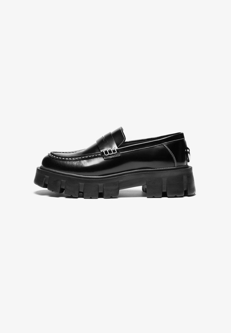Schwarze Lederloafer mit dicker Gummisohle, dekorativem Riemen über dem Obermaterial und kontrastierenden weißen Nähten. Glatte Textur mit abgerundeter Zehenpartie.