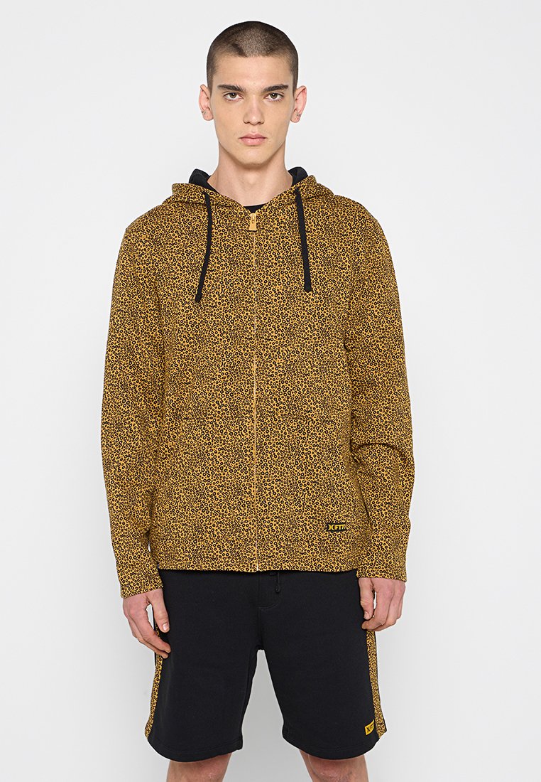 Kapuzensweatjacke mit einem gelb-schwarzen Leopardenmuster, ausgestattet mit einer Kängurutasche und schwarzen Kordeln. Kombiniert mit schwarzen Shorts.