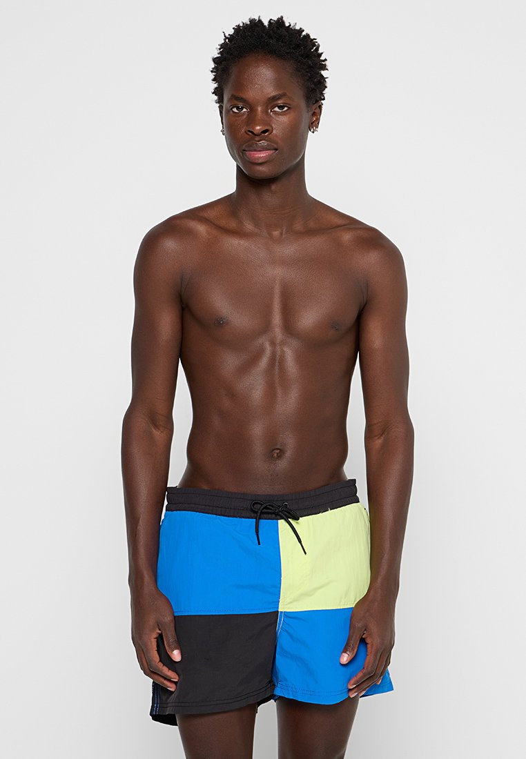 Herren-Badebermuda im Patchwork-Design mit blauen, grünen, schwarzen und gelben Paneelen. Hergestellt aus leichtem Stoff mit einem elastischen Bund.