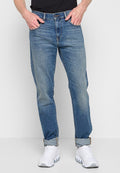 Mann in blauen hochgekrempelten Jeans und weißen klobigen Sneakern, Hände teilweise in den Taschen, vor einem schlichten hellen Hintergrund.
