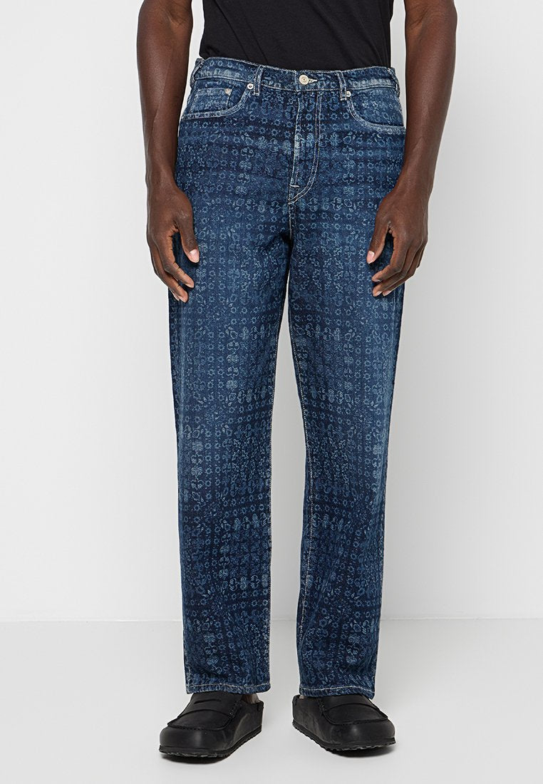 Mann in dunkelblauen, gemusterten, geraden Jeans, schwarzen Slippers und einem schwarzen Hemd, steht vor einem schlichten, hellen Hintergrund.