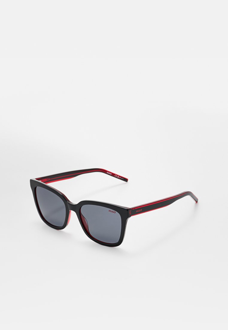 Schwarze rechteckige Sonnenbrille mit dunklen Gläsern und roten Akzenten im inneren Rahmen, mit dem Markennamen „HUGO“ auf Bügeln und Gläsern, vor weißem Hintergrund.