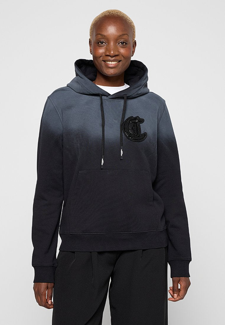 Lächelnde Person mit kurzen blonden Haaren, die einen Hoodie in Schwarz-Grau-Gradient mit einer Fronttasche und einem großen "C"-Logo trägt.
