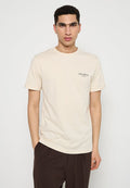 Junger Mann mit kurzen, lockigen Haaren, der ein cremefarbenes T-Shirt mit dem Aufdruck "LES DEUX" und dunklen Hosen trägt, steht mit einer Hand in der Tasche.