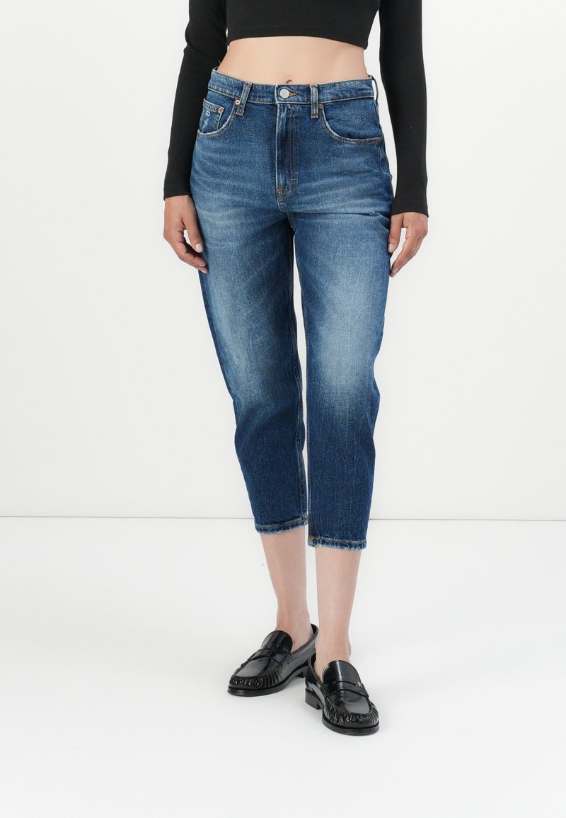 Hoch taillierte blaue Jeans aus Denim mit verkürzten Beinen, ausgewaschenen Stellen und umgeschlagenen Säumen. Kombiniert mit schwarzen Slippern und einem enganliegenden schwarzen Oberteil.
