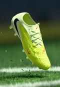 Helles gelbes Fußballschuh mit strukturierter Oberfläche, weißer Sohle, schwarzen und roten Akzenten, schwebt über grünem Gras mit Turfpartikeln.