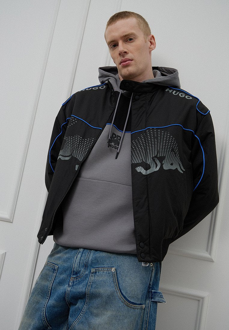 Schwarze Jacke mit blauen Akzenten und Logo, grauer Hoodie darunter und mittelblaue Jeans. Strukturierte Designelemente auf der Vorderseite der Jacke.