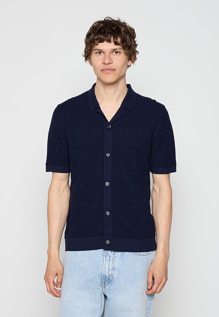 Junger Mann, der ein dunkelblaues, strukturiertes Kurzarmhemd mit Kragen und hellblaue Jeans trägt, vor einem einfarbigen Hintergrund steht.