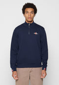 Junger Mann, der nach vorne blickt, trägt einen navyblauen Quarter-Zip-Sweater mit einem kleinen rot-weißen Logo auf Brust und Ärmeln sowie beigen Hosen.