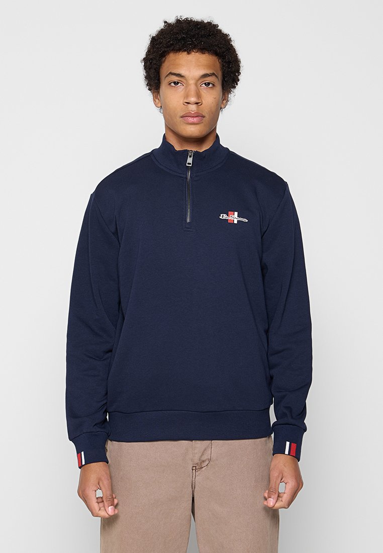 Junger Mann, der nach vorne blickt, trägt einen navyblauen Quarter-Zip-Sweater mit einem kleinen rot-weißen Logo auf Brust und Ärmeln sowie beigen Hosen.
