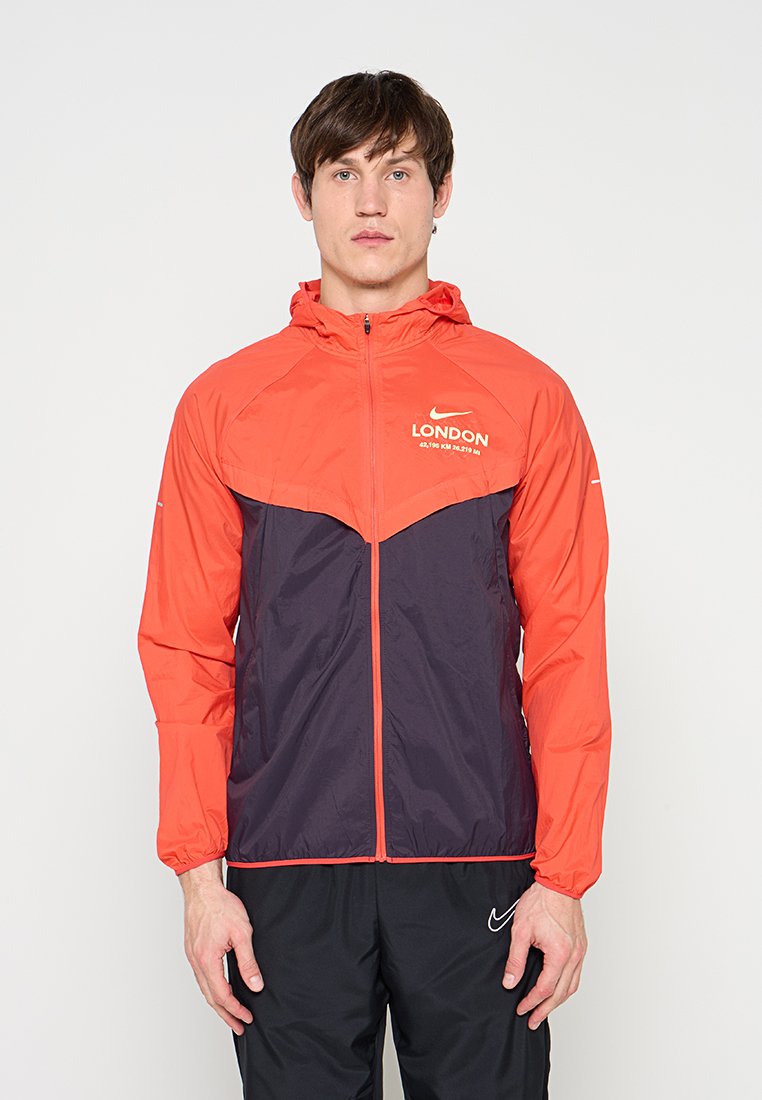 Mann trägt eine rote und schwarze Nike London Marathon Jacke mit Kapuze und schwarze Nike-Hose, steht vor einem schlichten weißen Hintergrund.